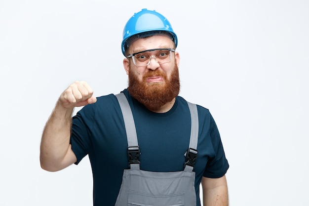 Unpleased young male construction worker wearing safety helmet uniform and safety glasses looking at camera keeping fist in air isolated on white background