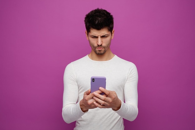 Unpleased young man using his mobile phone isolated on purple background