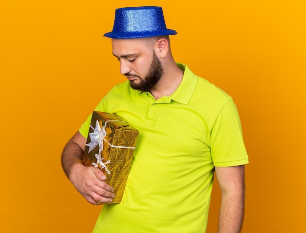 Unpleased young man wearing party hat holding and looking at gift box isolated on orange wall