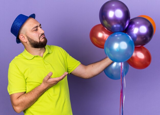 Unpleased young man wearing party hat holding and points at balloons 