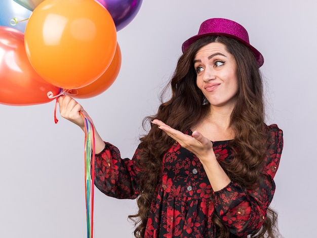 Una giovane ragazza scontenta che indossa un cappello da festa che tiene guardando e indicando con la mano i palloncini isolati sul muro bianco