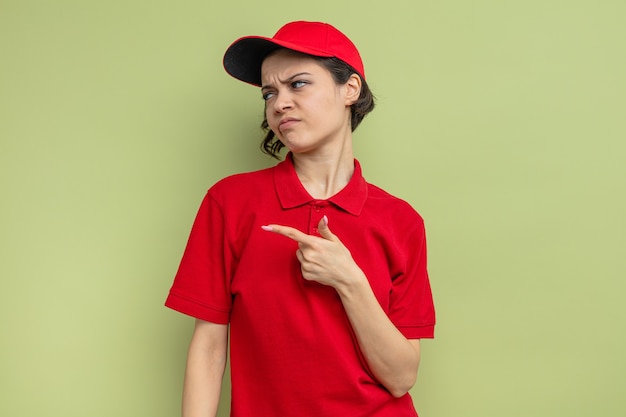 Unpleased young pretty delivery woman looking and pointing at side 