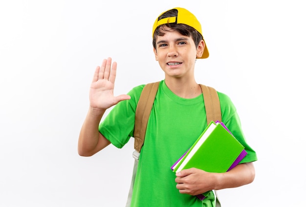 Unpleased young school boy wearing backpack with cap holding books showing stop gesture