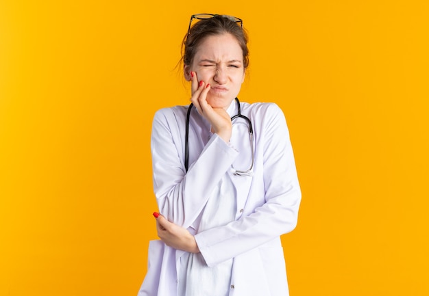 Unpleased young slavic girl in doctor uniform with stethoscope putting hand on her face and  isolated on orange wall with copy space