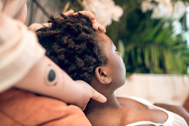 Unplugged. Caucasian hands with tattoo touching head and neck of lying dark-skinned woman with short curly hair in spa