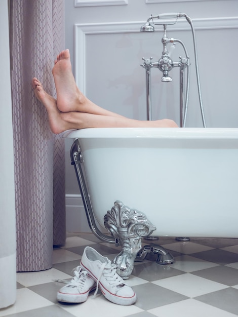 Foto donna irriconoscibile a piedi nudi che riposa nella vasca da bagno bianca con le gambe fuori dall'acqua e scarpe da ginnastica spente nel bagno luminoso