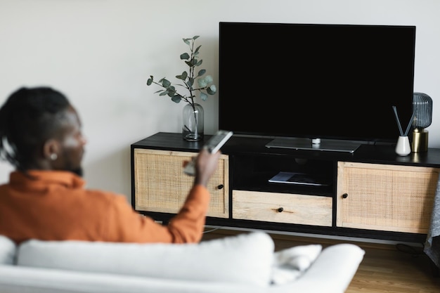 Unrecognizable black man turning on plasma flatscreen TVset pointing remote control at empty black TV screen mockup