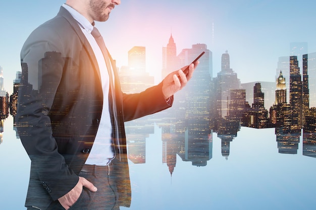 Неузнаваемый бизнесмен в костюме смотрит на смартфон на фоне городского пейзажа и его отражение. Двойная экспозиция тонированного изображения
