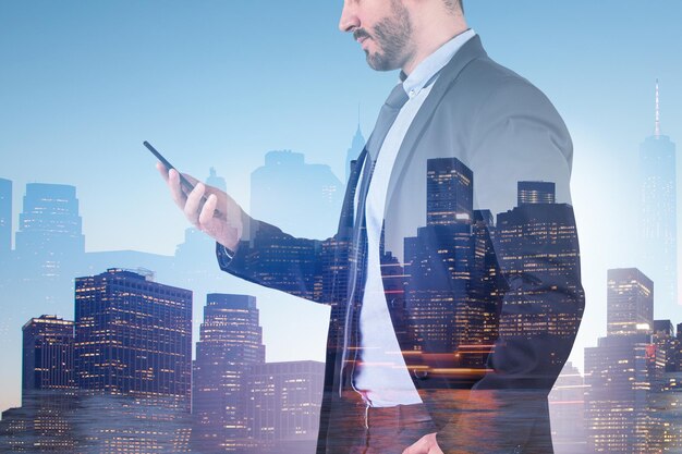 Unrecognizable businessman in suit looking at smartphone over night cityscape background with blue sky. Toned image double exposure