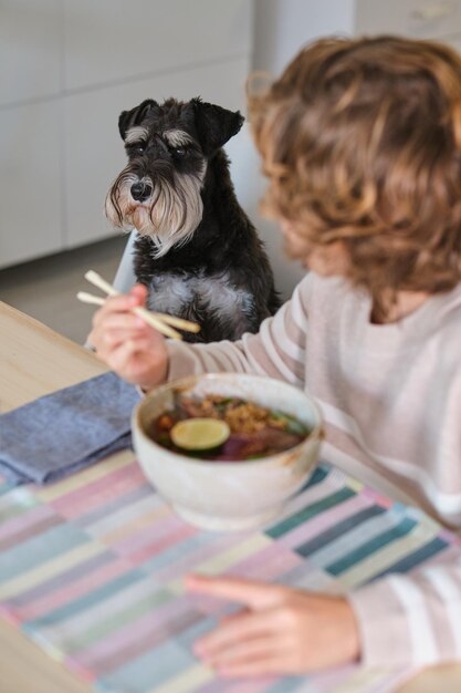 Unrecognizable child eating delicious soup with noodles using chopsticks while sitting at wooden table near pet in kitchen at home