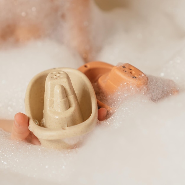 Unrecognizable child holding together cute beige and orange small silicone toy boats with holes on top in bubble bath