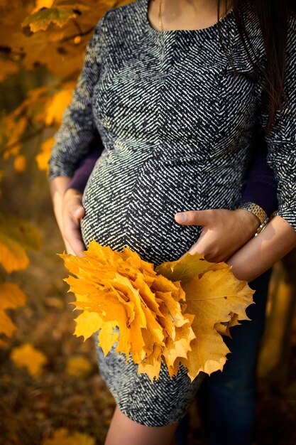 認識できないカップル、妊娠中の女性と彼女の夫は自然に腹を抱き、紅葉の間の森に立って、秋になります。新しい人生のコンセプト。