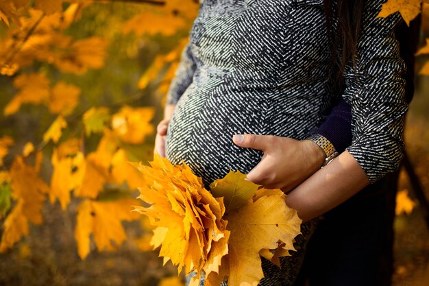 Unrecognizable  Couple, pregnant woman and her husband hugging belly on nature,  standing in forest among autumn leaves, fall. New life concept.