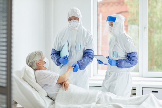 Unrecognizable doctors wearing protective suits with gloves, masks and eyewear working with patient in infectious disease ward