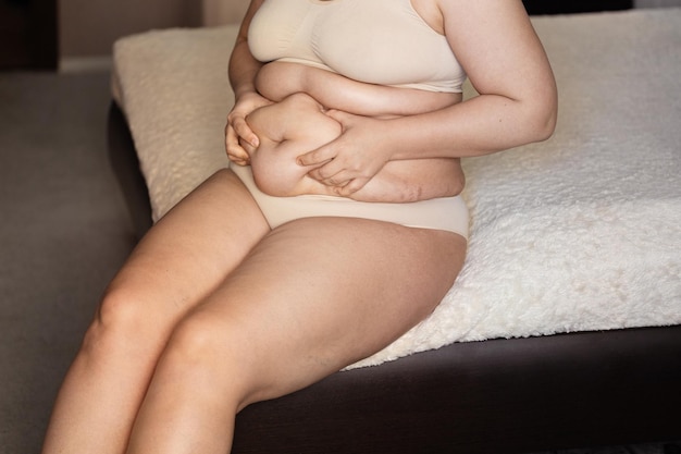 Unrecognizable fat woman sitting on edge of beige plaid in nude\
bra underpants showing squeezing excess naked belly