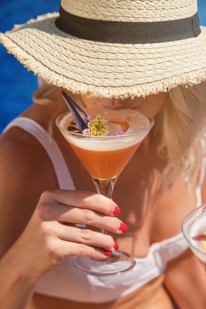 Unrecognizable female in straw hat and white swimsuit drinking refreshing cocktail while resting during summer day