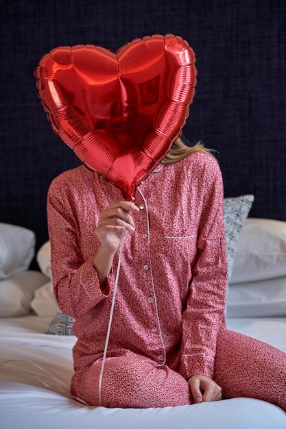 Unrecognizable female wearing stylish nightwear sitting on bed in hotel suite and hiding face behind gifted red heart shaped foil balloon