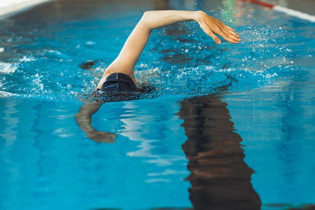 Unrecognizable fit professional female swimmer in cap swimming in pool during indoor training