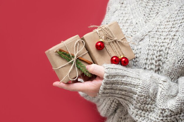 An unrecognizable girl in a sweater holds Christmas gifts on a red background. Handmade gift boxes in the hands of a woman. New year background with beautiful gifts.