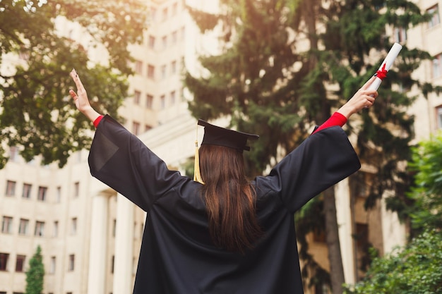 Unrecognizable happy woman on her graduation day at university holding diploma and raising hands, back view, copy space. Education, qualification and gown concept.