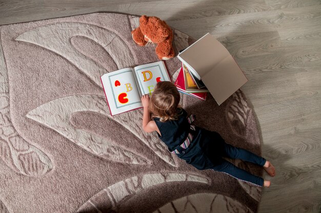 Unrecognizable little girl in a dark blue dress reading a book lying on the floor near teddy bear.