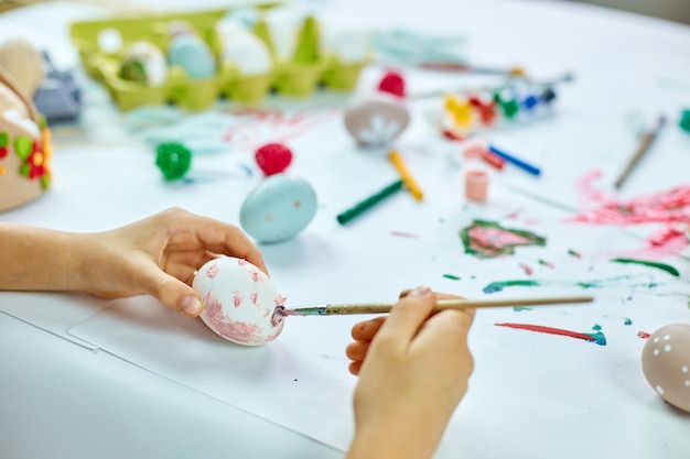 Unrecognizable little girl painting, drawing with brush eggs at home. Child preparing for Easter, having fun and celebrating feast. Happy Easter, DIY