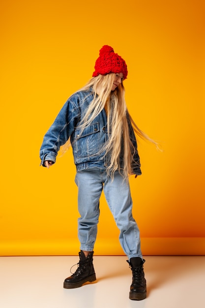 Modello femminile hipster dai capelli lunghi irriconoscibile che indossa giacca di jeans calda e jeans con cappello lavorato a maglia rosso in piedi su sfondo arancione in studio