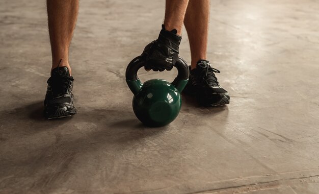 Unrecognizable male athlete grasping heavy kettlebell on concrete floor during weightlifting workout in gym