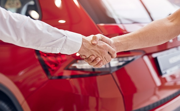 Фото Неузнаваемый мужчина-клиент и менеджер пожимают руку красному автомобилю в современном автосалоне