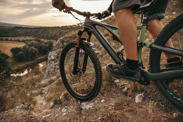 Ciclista maschio irriconoscibile che guida la bicicletta sul sentiero roccioso in terreno montuoso durante il tramonto in campagna