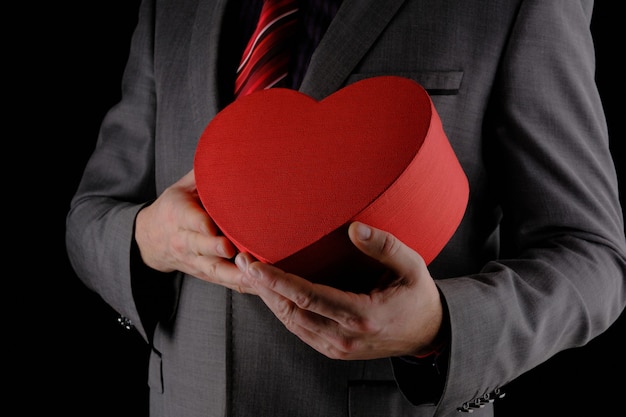 Unrecognizable man in gray suit holds out heart shaped gift red box, congratulatory concept, black background