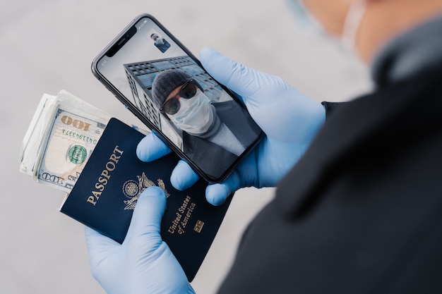 Unrecognizable man has video call, talks with friend, wears protective medical rubber gloves, holds passport with money, going to leave country during coronavirus pandemic. Social distance concept