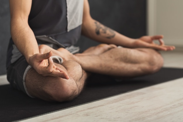 Unrecognizable man practicing yoga, meditation exercises. Guy does lotus pose for relaxation, sitting on mat at gym. Healthy lifestyle, crop