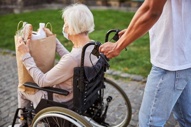 Unrecognizable man pushing the wheelchair outdoors while an aged lady holding a shopping bag