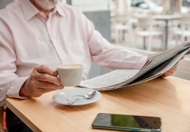 An unrecognizable man reading a daily newspaper holding up a cup of coffee with one hand the cell phone next to him on the wooden table