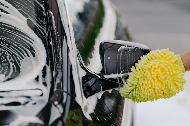 Unrecognizable man washes black car with cloth or rag mitten uses special detergent Cleaning dirty automobile Carwash business concept