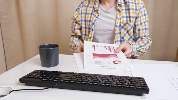 Unrecognizable man working with documents and typing text on the keyboard while drinking a drink from a cup.