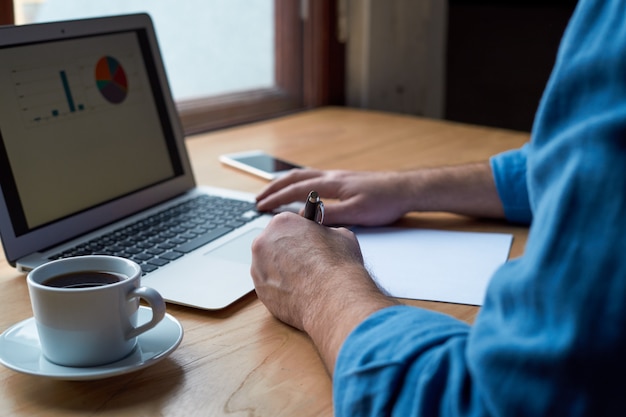 Unrecognizable man writes plan on paper and looks at charts on computer screen, laptop. 