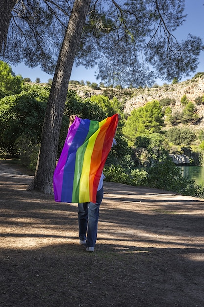 До неузнаваемости лесбиянка средних лет, держащая радужный гей-флаг на озере на открытом воздухе. Концепция свободы