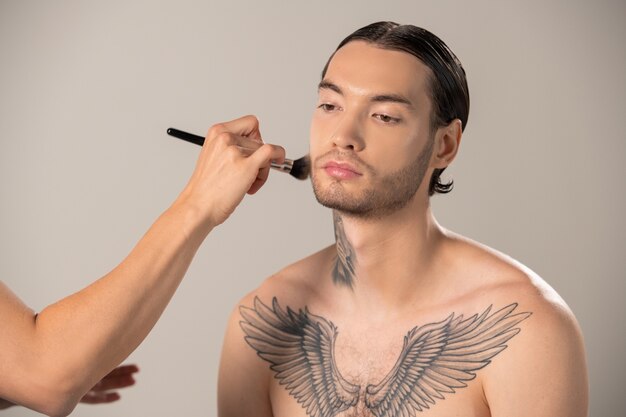 Unrecognizable MUA artist applying makeup to man with tattoo on chest in studio