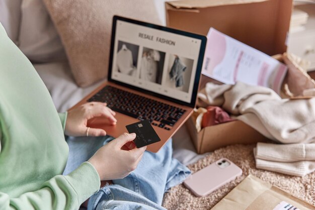 Photo unrecognizable person holds banking card and uses laptop computer makes shopping online buys new clothes on sale poses on bed reatail payment purchasing and shipping concept focus on credit card