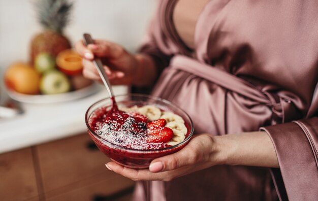 Unrecognizable pregnant female in silk robe eating porridge with strawberry and banana in morning at home