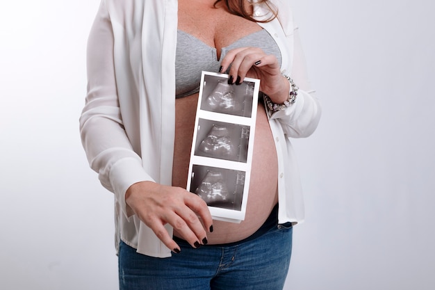 До неузнаваемости беременная женщина, держащая ультразвуковые изображения своего будущего ребенка над животом на белом фоне