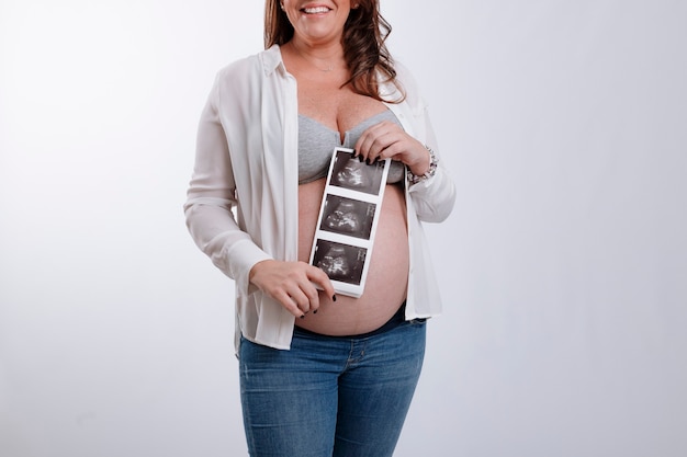 Фото До неузнаваемости беременная женщина, держащая ультразвуковые изображения своего будущего ребенка, улыбаясь на белом фоне