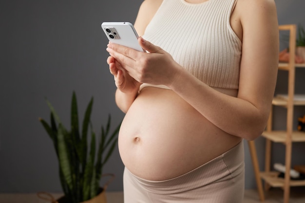 Фото Неузнаваемая беременная женщина с голым животом, стоящая в домашнем интерьере с смартфоном в руках, использующая мобильное приложение для будущих матерей, прокручиваясь или делая покупки в интернете