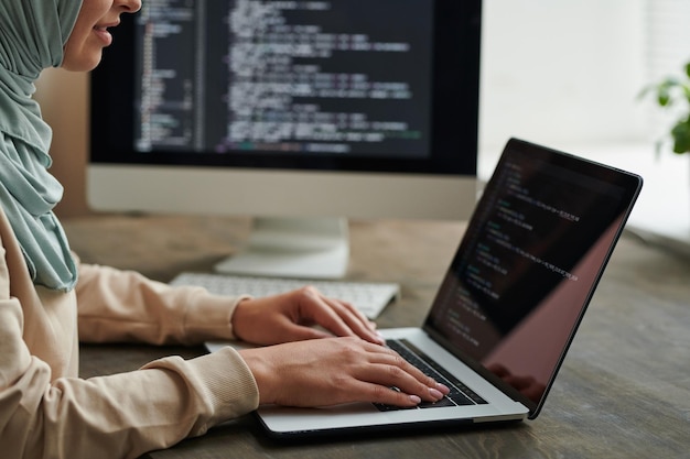 Unrecognizable professional programmer wearing hijab sitting at office table working on computer cod