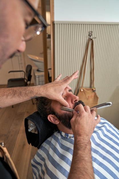 Unrecognizable serious barber in eyeglasses cutting beard of male client with straight blade in modern barbershop