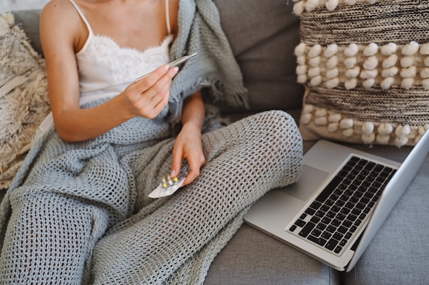 Unrecognizable sick woman on couch with laptop holding thermometer and pills, home quarantine self isolation. Online work from home. Corona virus infection COVID-19 concept.