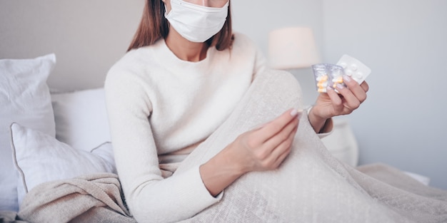 自宅検疫で温度計と薬を保持しているベッドでフェイスマスクで認識できない病気の女性