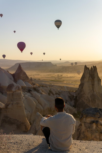 Unrecognizable tourist sitting in the foreground enjoying views of the valley of love and hot air balloons flying at sunrise in Cappadocia Turkey